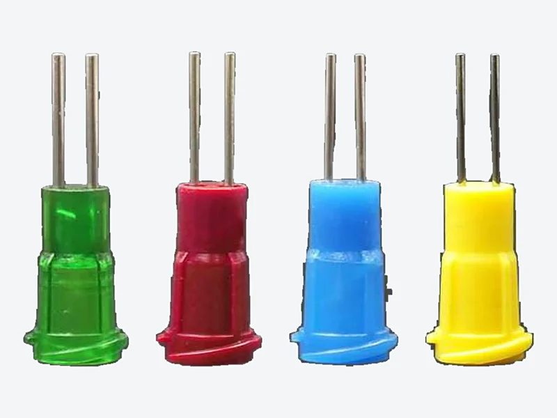 Four dispensing needles with green, dark red, blue and yellow plastic hubs, each with two parallel metal tubes